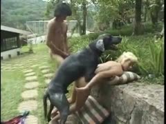 A sultry golden-haired person is bonked by a black dog in the street.